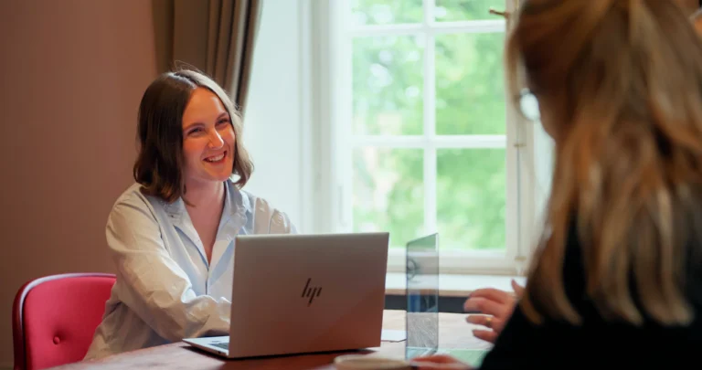 Kvinna ler mot kollega under möte, sitter vid bord med laptop nära fönster i ljust rum.