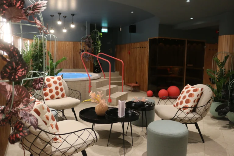 Modern spa-lounge med bubbelpool, trådramsstolar, prickiga kuddar, växter och röda accenter. Träpaneler och pendellampor skapar atmosfär.