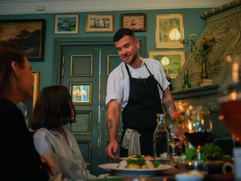 Kock i förkläde interagerar med gästerna vid bordet. Restauranginredning med turkosa väggar, inramade konstverk och stämningsbelysning.