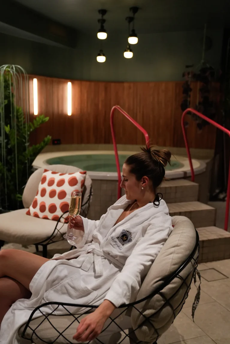 Kvinna i badrock kopplar av i spa-fåtölj och håller i ett champagneglas. Bubbelpool, stämningsbelysning och växter skapar en lyxig atmosfär.