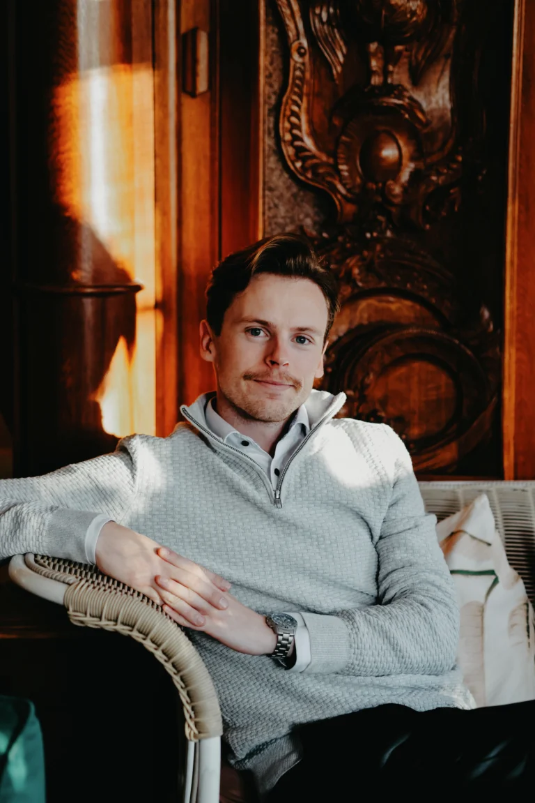 Man i grå tröja sitter i rottingstol, varm interiör med utsmyckad inredning och öppen spis synlig i bakgrunden.