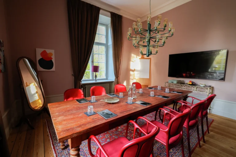 Elegant matsal med träbord, röda stolar, ljuskrona, rosa väggar, fönster med gardiner, stor spegel och vägghängd TV.