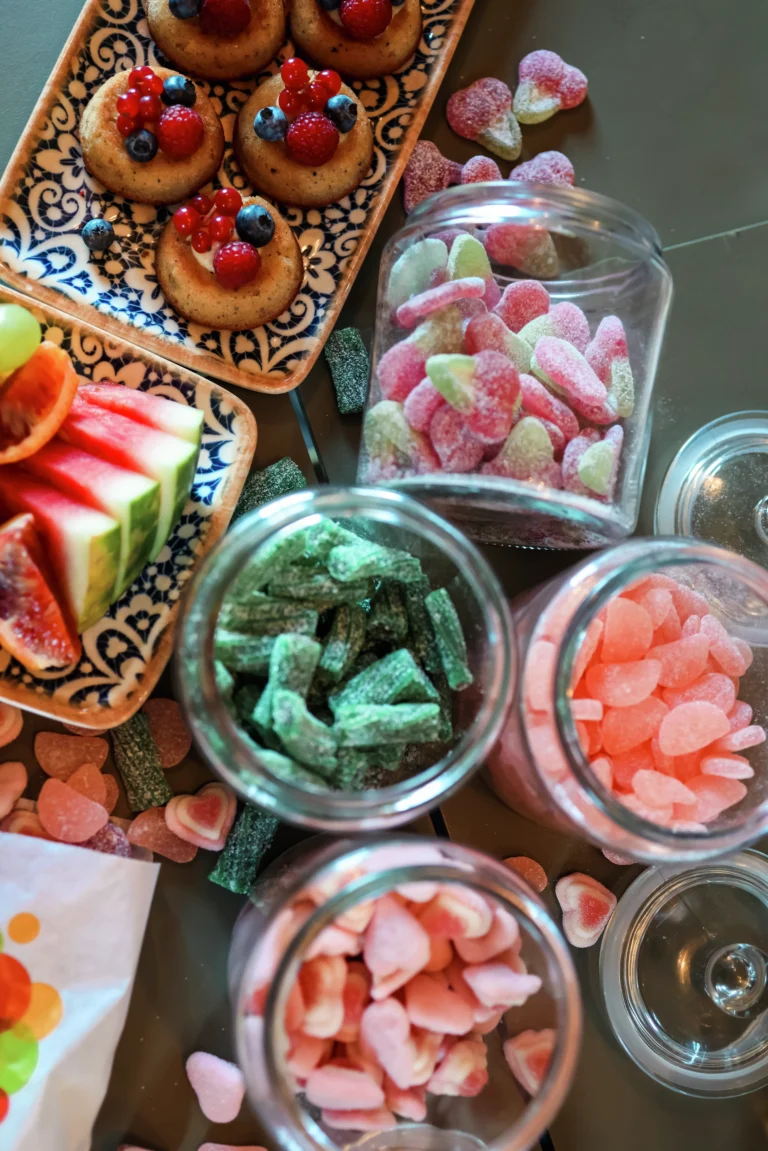 Blandade sötsaker och frukter på dekorativa tallrikar och i burkar, inklusive bakverk med bär, vattenmelon och färgglada gummigodisar.
