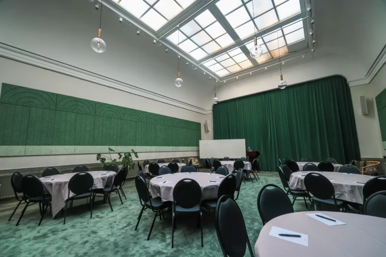 Konferensrum med takfönster, runda bord, grön matta och gardiner. Pendellampor och dekorativa väggpaneler skapar en elegant atmosfär.