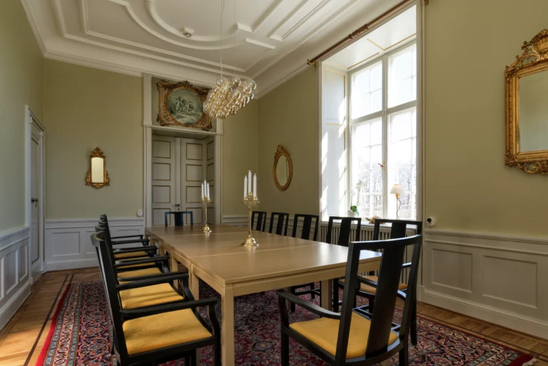 Elegant matsal med utsmyckat tak, stora fönster, antika speglar, ljuskrona, långt bord med svarta stolar och mönstrad matta på trägolv.