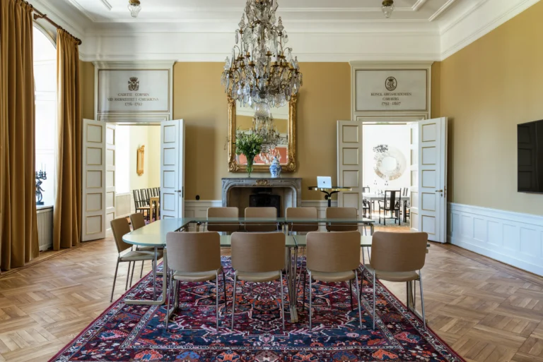 Elegant matsal med kristallkrona, utsmyckad spegel, modernt bord och stolar, persisk matta och dubbeldörrar som leder till angränsande rum.