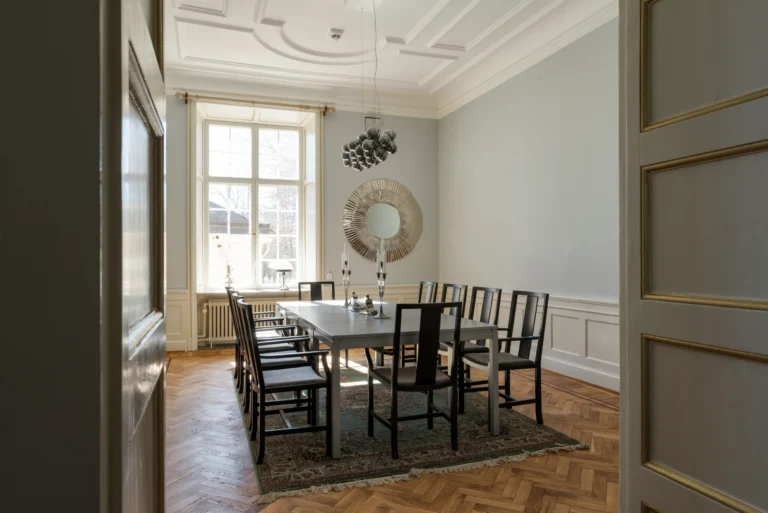 Elegant matsal med utsmyckat tak, stort fönster, modernt svart bord och stolar, rund spegel och fiskbensmönster.