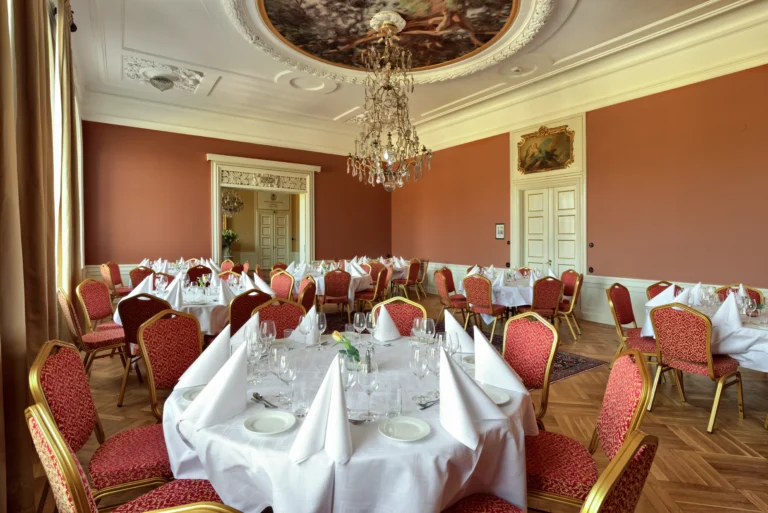 Elegant matsal med utsmyckat tak, kristallkrona, runda bord dukade för formell middag och rosa stoppade stolar på parkettgolv.