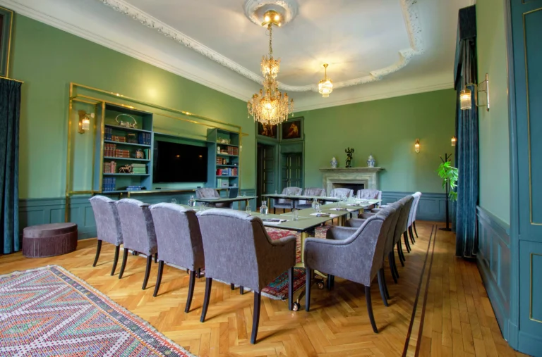 Elegant matsal med gröna väggar, kristallkrona, långt bord med lila stolar, bokhylla, öppen spis och fiskbensmönster.